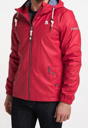 Homme portant une veste à capuche rouge avec des cordons blancs, une fermeture éclair et des poches latérales boutonnées, assortie d'un jean foncé.