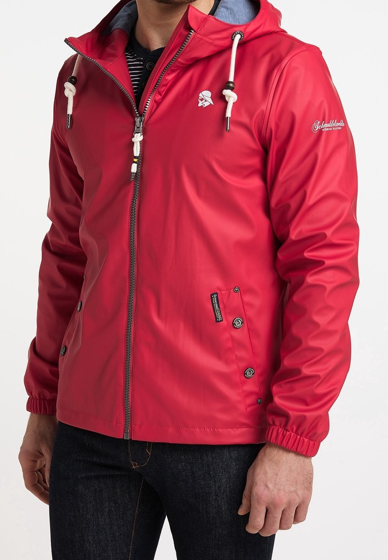 Homme portant une veste à capuche rouge avec des cordons blancs, une fermeture éclair et des poches latérales boutonnées, assortie d'un jean foncé.