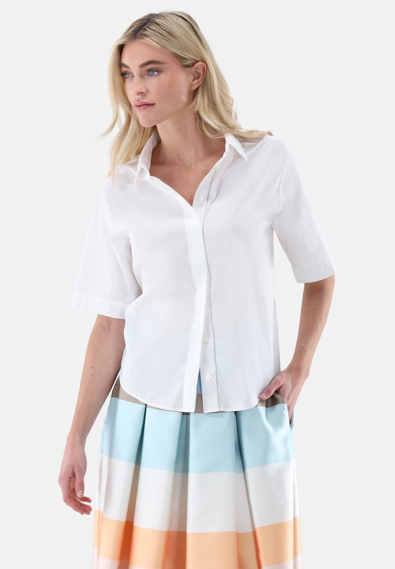 Short-sleeve white button-up shirt made of lightweight fabric with a classic collar and a straight hem. Layered over a pleated skirt.