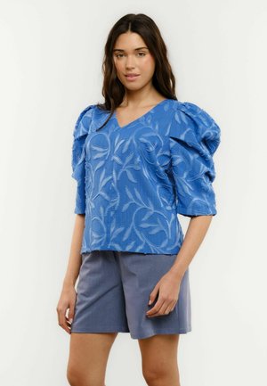 Nő kék, texturált virágmintás, puffos ujjú topban, V-nyakkal és szürke térdig érő rövidnadrágban, egyszínű háttér előtt állva.