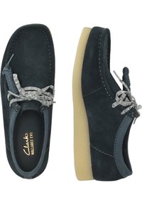 Tummansinisiä mokkanahkakenkiä, joissa on pyöreä kärki, ommeltu yksityiskohta ja harmaa nauhakiinnitys. Kevyt, vaalean kermanvärinen kumipohja. Clarks-logolla varustettu sisus.