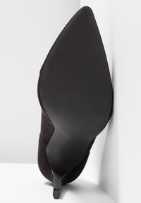 Chaussure noire à talons hauts avec bout pointu, dessus en daim, et talon aiguille fin. La semelle est lisse avec de légers motifs de traction.