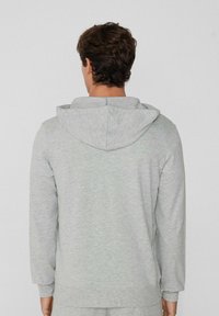 Mann in einer hellgrauen Langarmkapuzenjacke, der mit dem Rücken zur Kamera vor einem einfarbigen weißen Hintergrund steht.