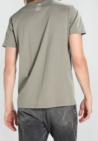 Camiseta de manga corta en verde oliva con cuello redondo y dobladillo recto. Textura ligera y suave. La parte posterior presenta una pequeña impresión de logo.
