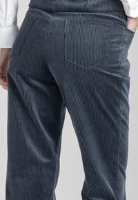 Osoba w ciemnoniebieskich spodniach sztruksowych, z rękami w kieszeniach, w połączeniu z białą koszulą z długim rękawem.