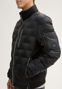 Černá prošívaná bunda s vpředu na zip, bočními kapsami a vysokým límcem; obsahuje jemné logo a kontrastní šití.