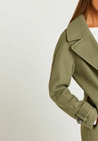 Donna che indossa un cappotto di lana verde oliva con ampie reverse e polsini abbottonati, posando con le mani in tasca su uno sfondo semplice.