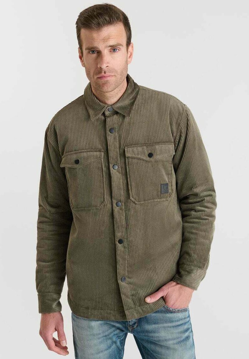 Man in olivegroene corduroy overhemd met knopen en klepzakken, staand met één hand in de zak van zijn blauwe jeans tegen een effen achtergrond.