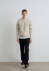 Μπεζ φούτερ με ανάγλυφο λογότυπο "BOSS", συνδυασμένο με άσπρη μπλούζα και μαύρο παντελόνι. Το μοντέλο στέκεται μπροστά σε ουδέτερο φόντο.