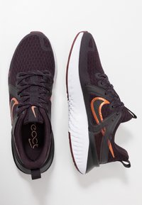 Nike React -lenkkarit, joissa tummanvioletti verkkomateriaali päällisosassa, mustia yksityiskohtia, oranssi logo ja valkoinen kuvioitu pohja harmaalla sisäosalla.