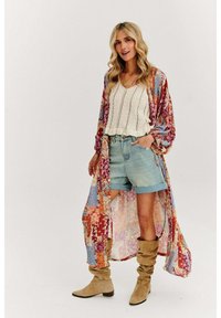 Dlhý, vzorovaný kimono s mnohobarevnými kvetinovými motívmi, nosené cez biele pletené tričko a svetlomodré džínsy s vysokým pásom, doplnené béžovými voľnými čižmami.