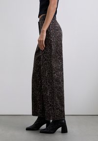 Woman wearing wide-leg leopard print pants with large side pocket and black block heel ankle boots on gray floor.