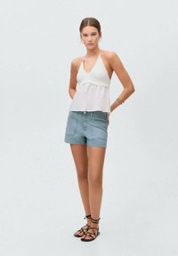 Bela halter majica s valovitim rubovima i svetloplave denims šortse. Manekenka nosi crne ravne sandale sa trakama, dok stoji ispred svetle pozadine.