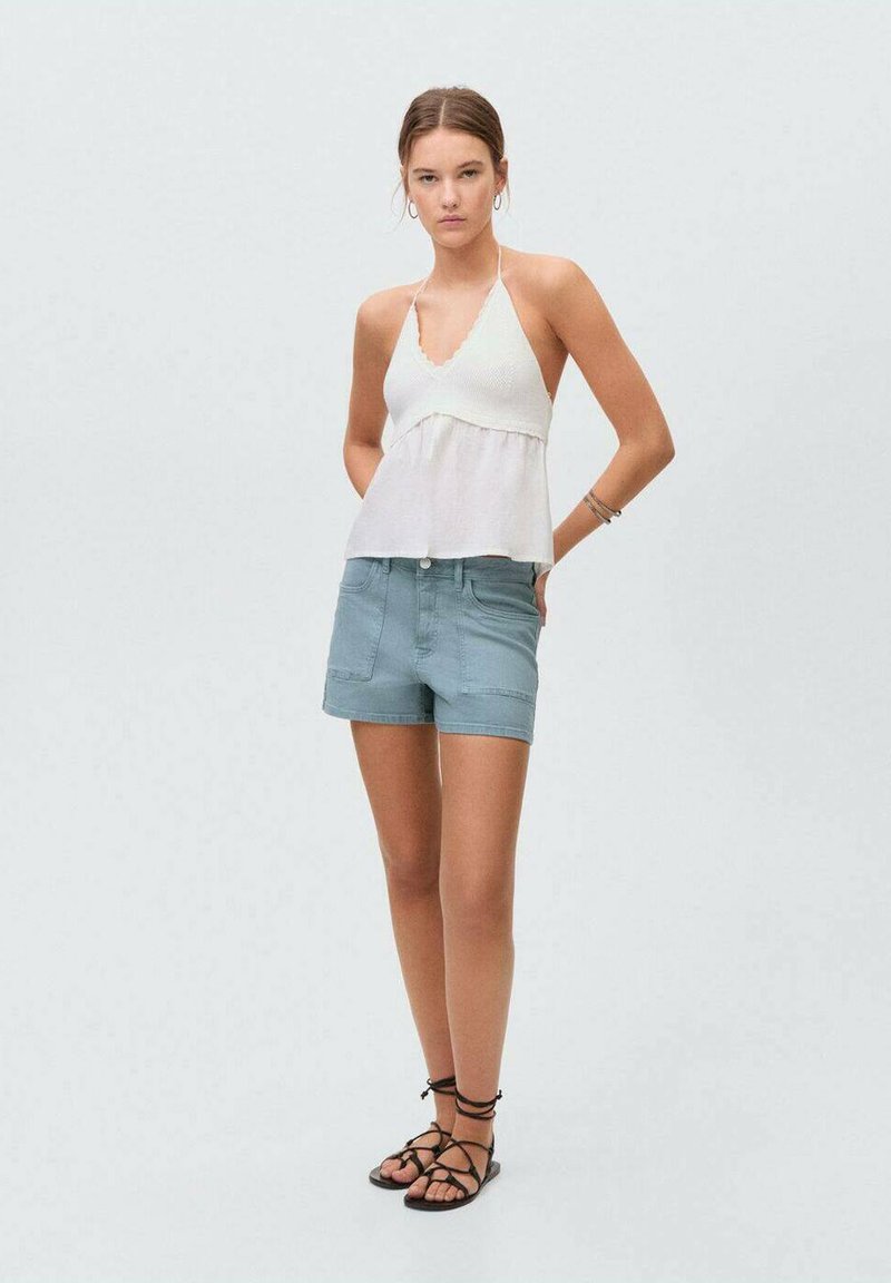 Bela halter majica s valovitim rubovima i svetloplave denims šortse. Manekenka nosi crne ravne sandale sa trakama, dok stoji ispred svetle pozadine.