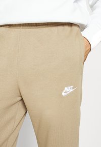 Bežové Nike tepláky vyrobené z mäkkej zmesi bavlny s elastickým pásom a bielym logom na ľavom stehne. Hladká textúra, jednoduchý dizajn.