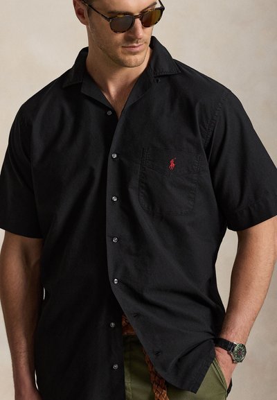 Man draagt een zwart overhemd met korte mouwen en een rood logo op de zak, een zonnebril, olijfgroene broek, een bruine gevlochten riem en een polshorloge.