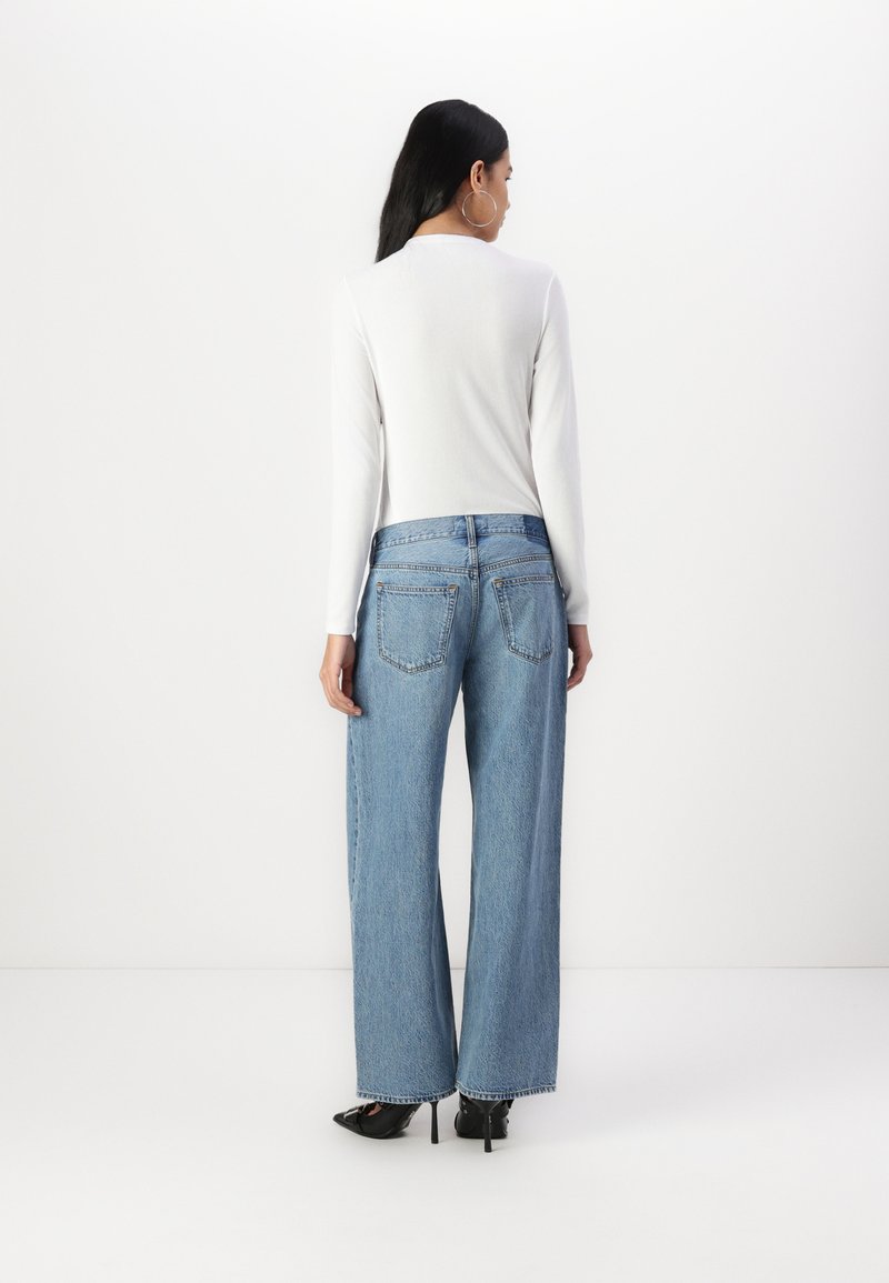Blaue weite Jeans mit hoher Taille, die klassische Gesäßtaschen und einen rohen Saum aufweisen, kombiniert mit einem weißen Langarmoberteil.
