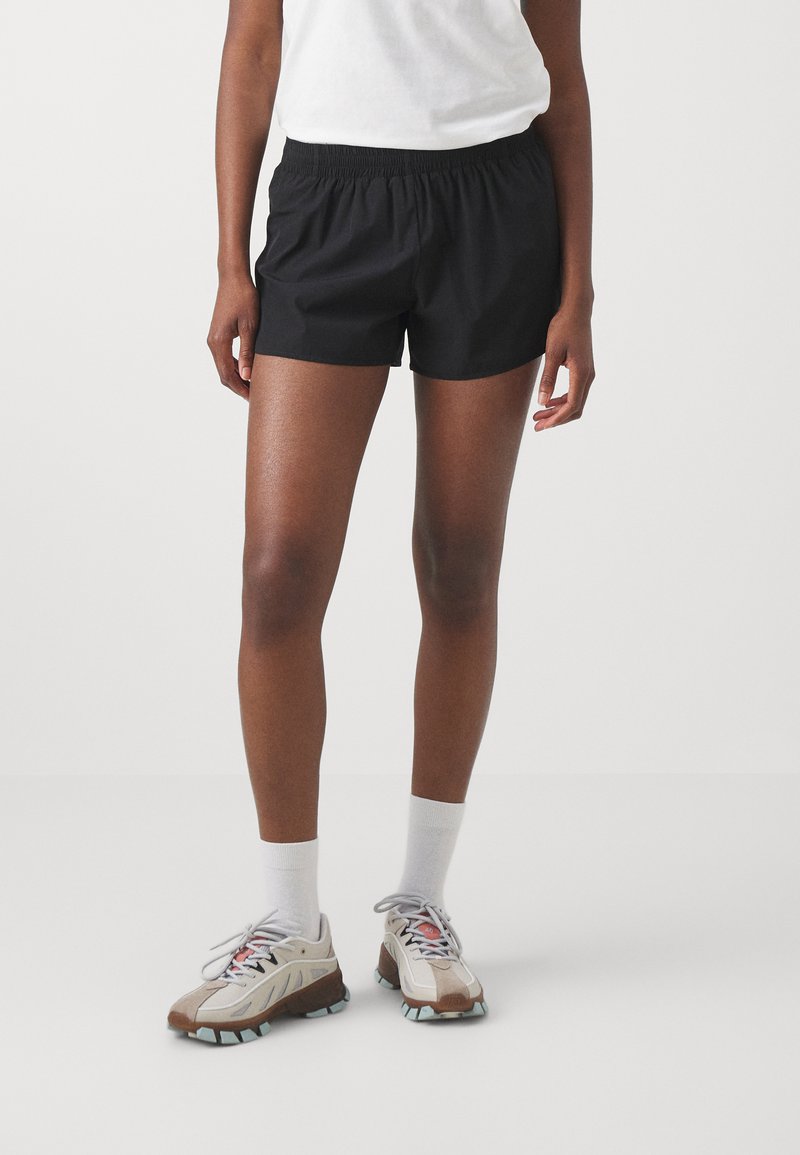 Svart idrettsshorts med elastisk midje, laget av lett materiale. Kombinert med beige og grå joggesko, og hvite ankelsokker.