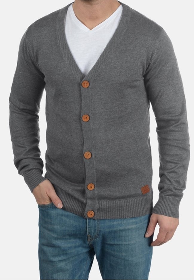 Homme portant un cardigan gris boutonné sur un t-shirt blanc à col en V et un jean bleu, une main dans la poche et l'autre le long du corps.