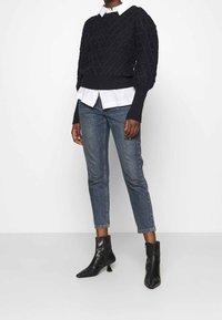 Svart stickad tröja med texturerat mönster, lager-på-lager över en vit skjorta med krage, parat med blå jeans i ankellängd och svarta ankle boots.