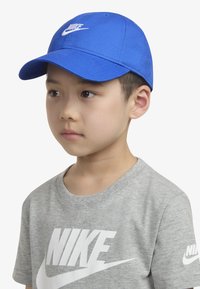 Blauwe Nike pet met gebogen klep en geborduurd logo. Grijs Nike T-shirt met grote witte logo-tekst. Strak ontwerp en zachte textuur.