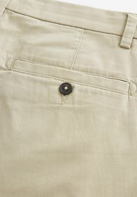 Pantalones de algodón beige con una textura suave, que cuentan con un bolsillo trasero asegurado por un botón oscuro y detalles de costura a medida.