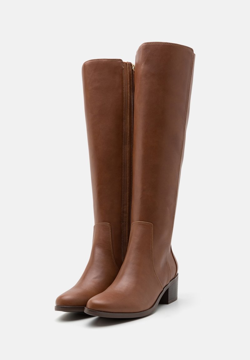 Bottes en cuir marron montantes jusqu’au genou, avec un bout rond et un talon en bloc. Elles présentent une fermeture éclair sur le côté et une texture lisse, rehaussée de coutures visibles.