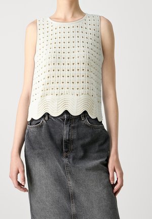 Kobieta w białym, bezrękawnikowym dzianinowym topie z falistym brzegiem i ciemnoszarej, wysokowariantowej spódnicy dżinsowej, ręce swobodnie opuszczone wzdłuż ciała.