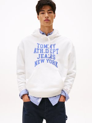 Felpa bianca con testo blu che recita "TOMMY ATH. DEPT JEANS NEW YORK," sovrapposta a una camicia a quadri blu e pantaloni scuri. Design semplice.