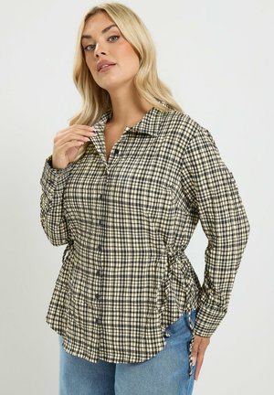 Blondine vrouw met een zwart-beige geruit, langärmelig overhemd met kraag en blauwe spijkerbroek tegen een effen achtergrond.