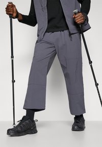 Gray outdoor pants made from lightweight material, featuring a relaxed fit, subtle texture, and practical utility pockets. Black hiking shoes visible.