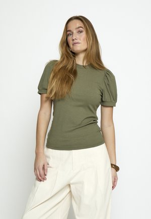 Žena s dugom kosom koja nosi maslinasto zelenu kratku rebrastu majicu i široke bijele hlače, stojeći ispred jednobojne bijele pozadine.