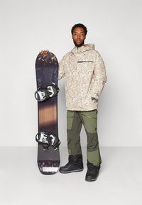 Snowboard avec un design à dégradé sombre, des fixations noires, tenu par une personne portant une veste en camouflage beige et un pantalon vert. Bottes noires.