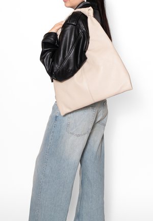 Borsa tote in pelle beige dalla forma triangolare, con una texture morbida, un design morbido e una tracolla singola, indossata sopra una giacca nera.