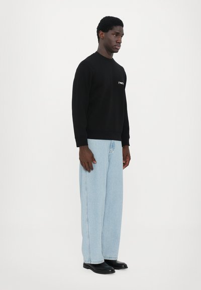 Sweatshirt noir avec logo sur la poitrine gauche, associé à un jean bleu clair à jambes larges. La tenue présente un design épuré et minimaliste.