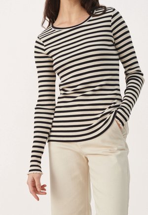 Femme portant un haut à rayures horizontales noir et blanc à manches longues et un pantalon beige clair, avec une main dans la poche, devant un fond uni.