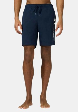 Uomo che indossa pantaloncini blu navy a metà coscia con un logo Champion bianco sulla gamba sinistra, in piedi a piedi nudi su uno sfondo semplice.
