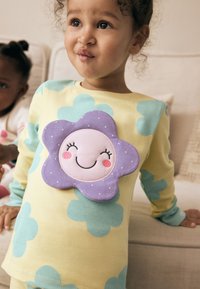 Haut à manches longues jaune avec de grands motifs de fleurs bleues et une application en peluche violette en forme de fleur avec un visage souriant et des joues roses.