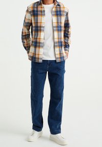 Chemise à carreaux boutonnée en marine, orange et crème ; t-shirt blanc en dessous ; associée à un jean bleu foncé et des baskets blanches.