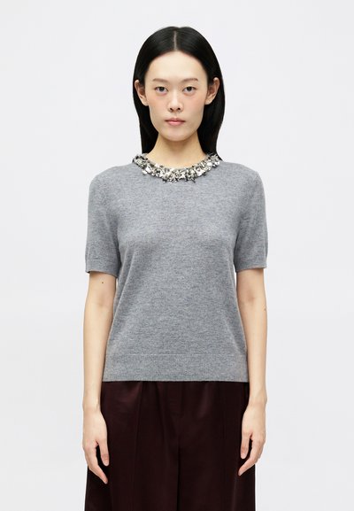 Un pull gris à manches courtes présente un col orné de perles et une texture douce, associé à un pantalon large sombre et taillé.
