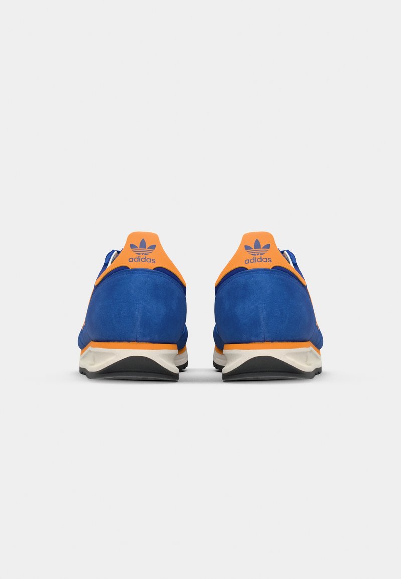 Zapatillas deportivas de gamuza azul con detalles en naranja y suelas blancas. Las características incluyen un talón redondeado y un logotipo en la parte trasera. Textura suave en general.