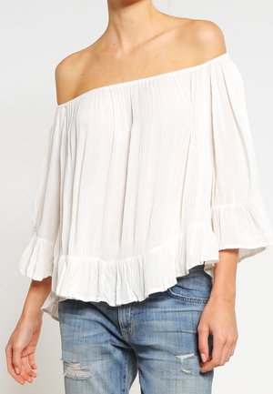 Blusa bianca a spalle scoperte con trama plissettata e orlo a volant. Maniche fluide e tessuto leggero. Abbinata a jeans strappati blu chiaro.