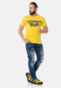 Yellow graphic t-shirt with blue and black design; blue distressed jeans with multiple pockets; yellow and black sneakers with textured soles.