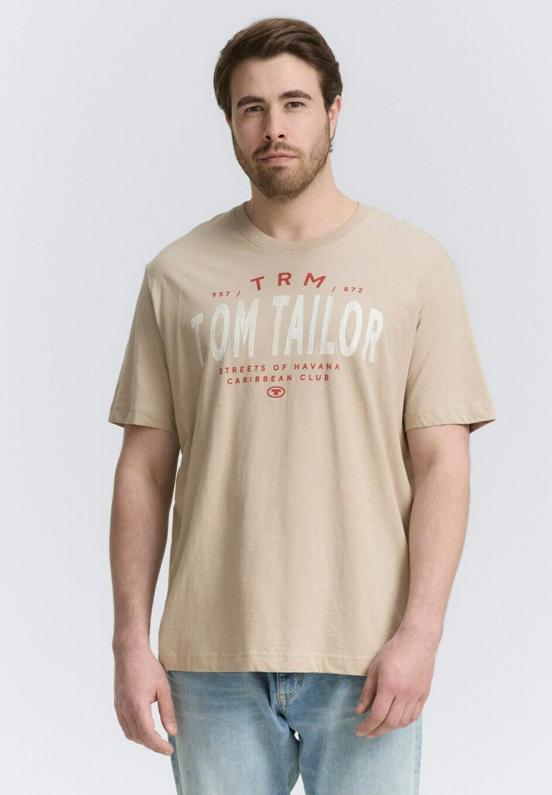 Beige Baumwoll-T-Shirt mit grafischem Text in Rot, lässiger Schnitt, kurze Ärmel und gerippter Rundhalsausschnitt, kombiniert mit hellblauen Jeans.