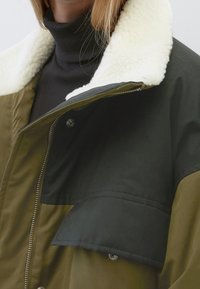 Olivgrüne Jacke mit einem schwarzen oberen Abschnitt, ausgestattet mit einem weichen, weißen Schafsfellkragen und einem Reißverschluss. Enthält eine Brusttasche und Metalldruckknöpfe.