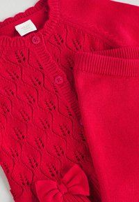 Roter gestrickter Cardigan mit einem Spitzenmuster, der pinke Knöpfe, gerippte Kanten und eine Schleifenakzent auf der Vorderseite aufweist. Glatte Textiloberfläche.