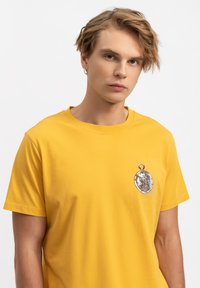 T-shirt di cotone giallo con scollo rotondo e maniche corte. Presenta un design a charm in tonalità argento sul lato sinistro del petto. Texture morbida.