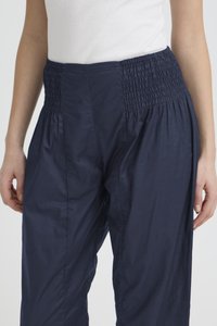 Moteris vilki tamsiai mėlynas aukštu liemeniu kelnes su plačia elastinga raukšlėta juosmens dalimi ir baltą be rankovių ribinę palaidinę.