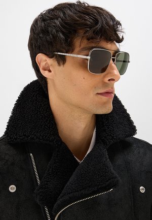 Uomo che indossa occhiali da sole quadrati scuri e una giacca in shearling nera con collo largo, guarda verso destra su uno sfondo uniforme.