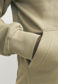 Beige sweatshirt met geribbelde manchetten en een borstzak. Zachte, gestructureerde stof toont stikdetails. Close-up van een hand in de zak.